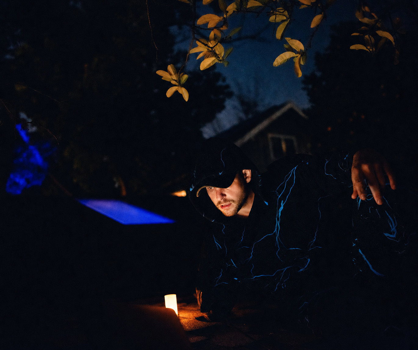 Cody Tarpley photographed at night in a hooded jacket, lit by a single candle on the pavement.